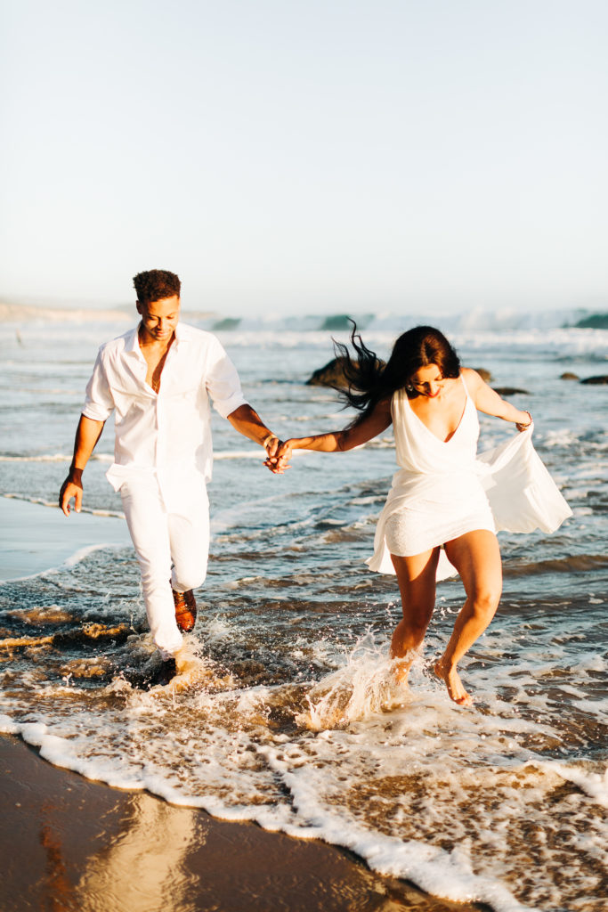 southern california engagement photos locations; couple holding hands and running through the waves at crystal cove state beach