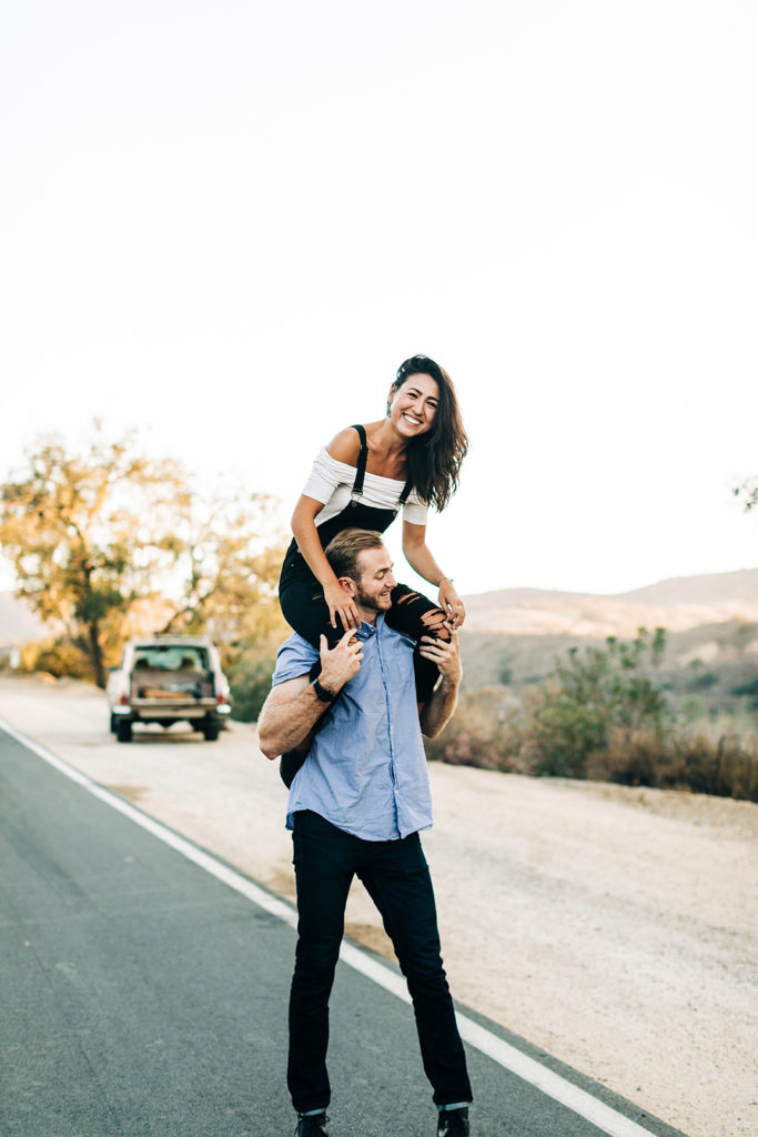 southern california engagement photos; woman sitting on man's shoulders while smiling on the side of the road with a vintage jeep in the background