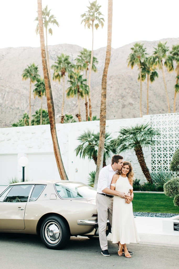 southern california engagement photos; couple hugging while standing next to a vintage car and palm trees in palm springs