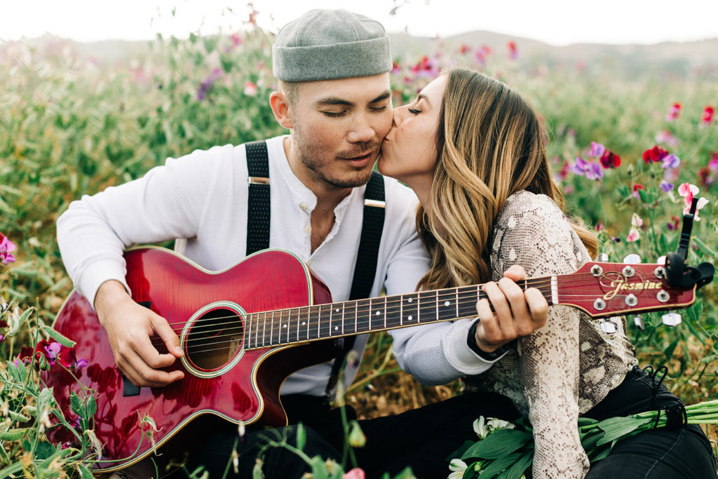 orange county engagement photos; man plays guitar for woman kissing him on the cheek while sitting in a field of flowers