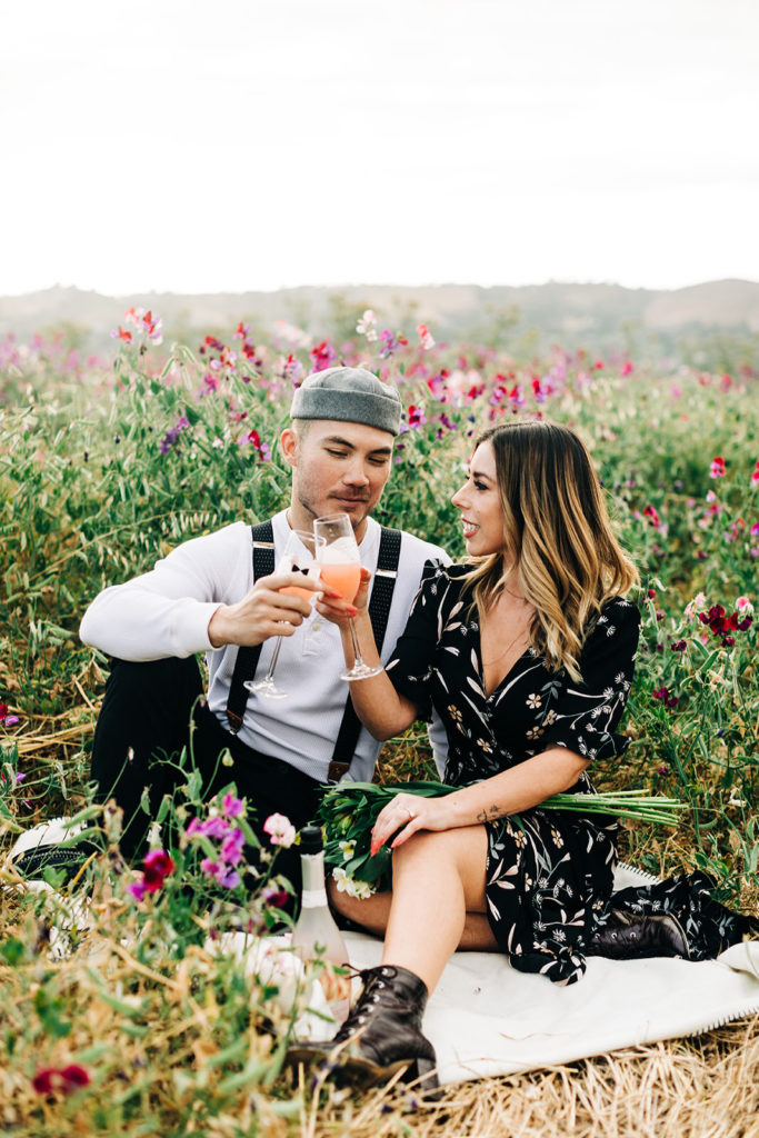 orange county engagement photos; man and woman smile while holding champagne in a field of flowers