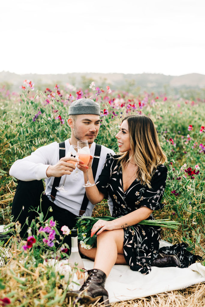 orange county engagement photos; man and woman smile while holding champagne in a field of flowers