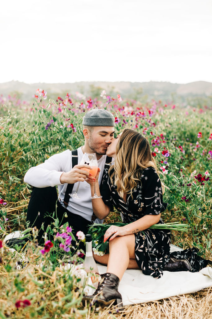 orange county engagement photos; man and woman kiss while holding champagne in a field of flowers
