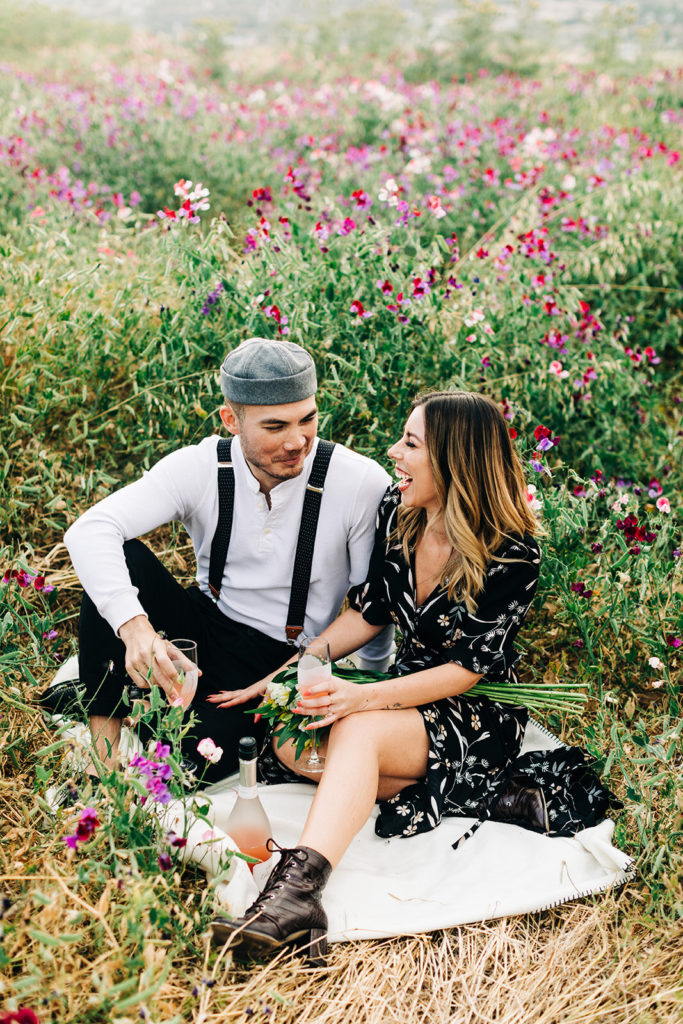 orange county engagement photos; couple sitting in a field of flowers and laughing