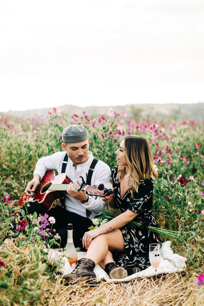 orange county engagement photos; man plays guitar for woman while sitting in a field of flowers