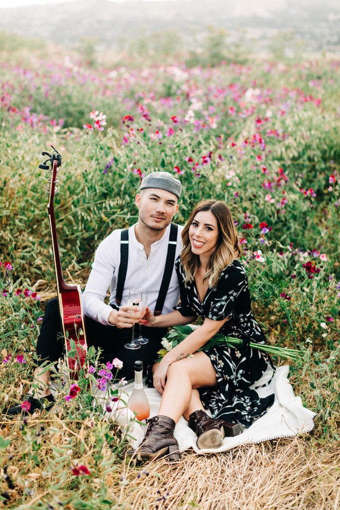 orange county engagement photos; couple sitting in a flower field with champagne and a guitar
