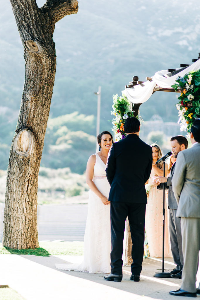 Sweet Pea Ranch In Upland, CA wedding photography; brise and groom listening to the vows in front of a tree and beautiful mountain view in the background
