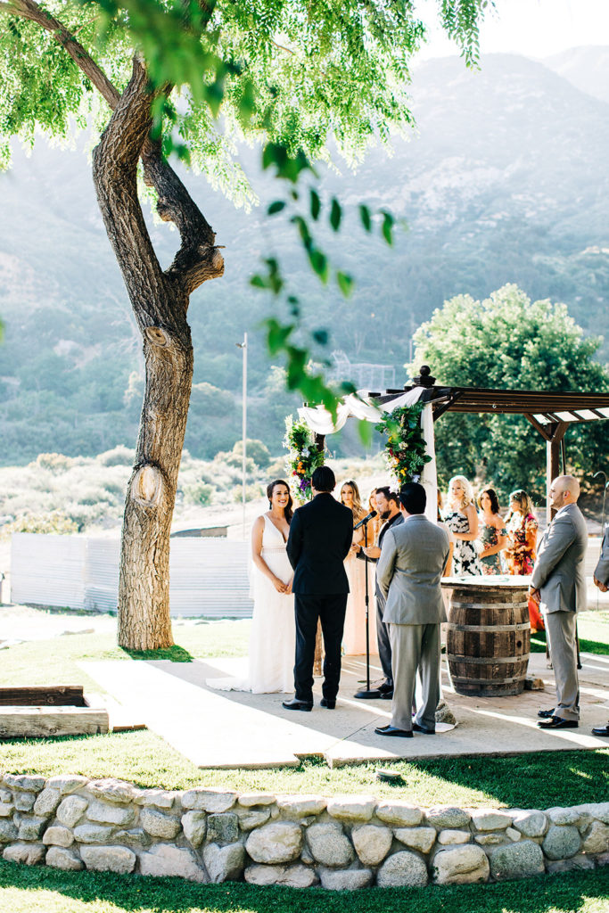 Sweet Pea Ranch In Upland, CA wedding photography; bride and groom listening to the vows in front of tree and beautiful mountain