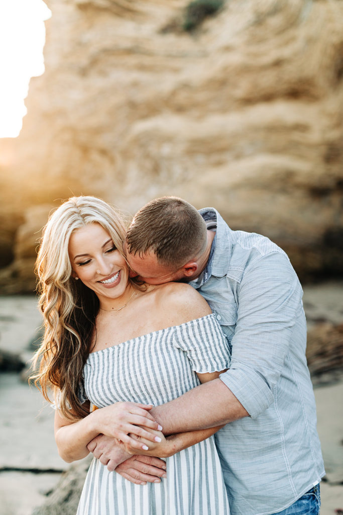 crystal cove beach engagement photos in orange county; man kissing woman on the neck while hugging her from behind