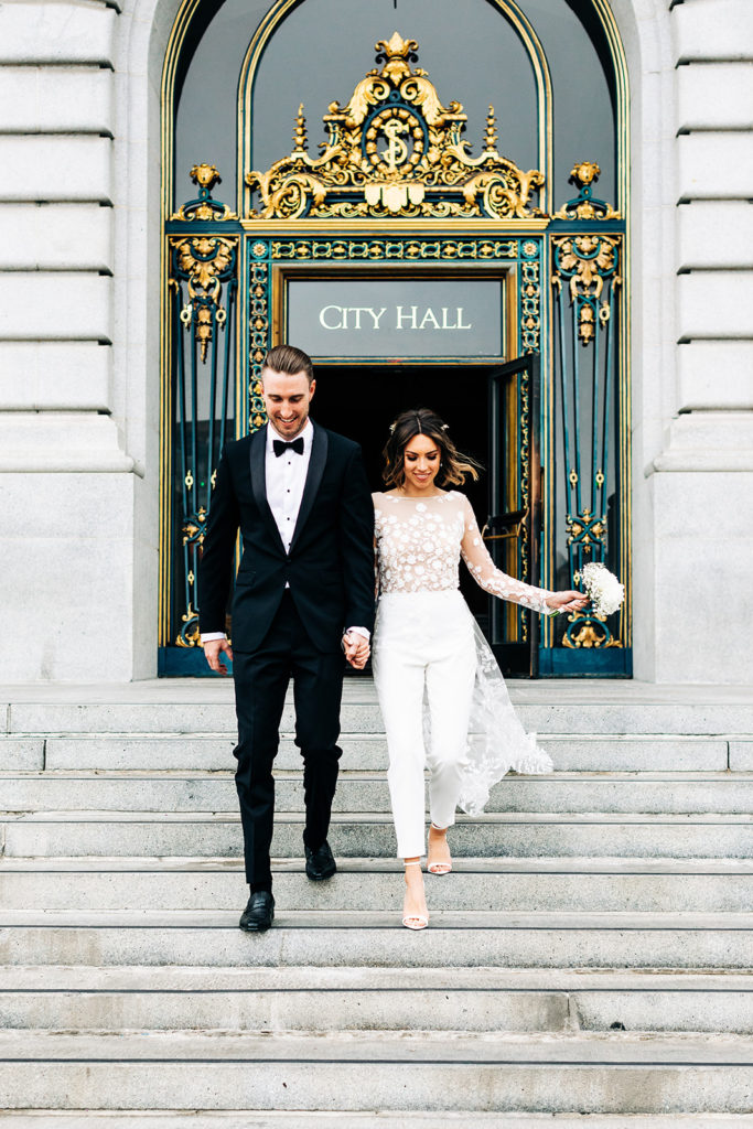 san franciso city hall elopement, san diego wedding photographer; bride and groom holding hands and running down cement steps, the words "city hall" are on the building behind them