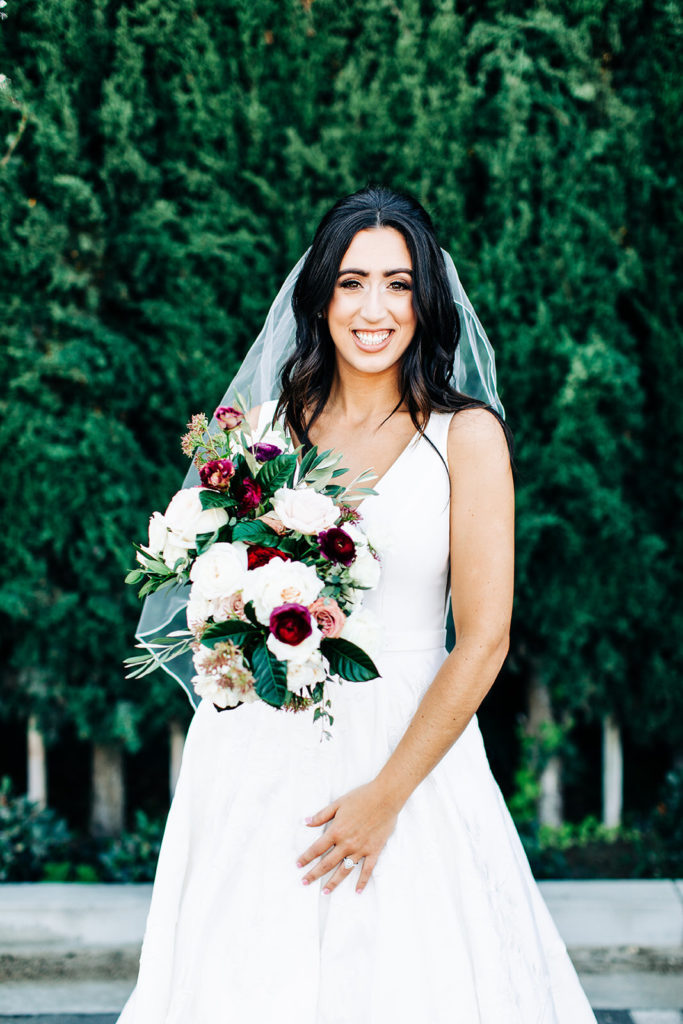 Colony house in Anaheim, CA wedding photography; bride posing in front of green trees with flowers in hand