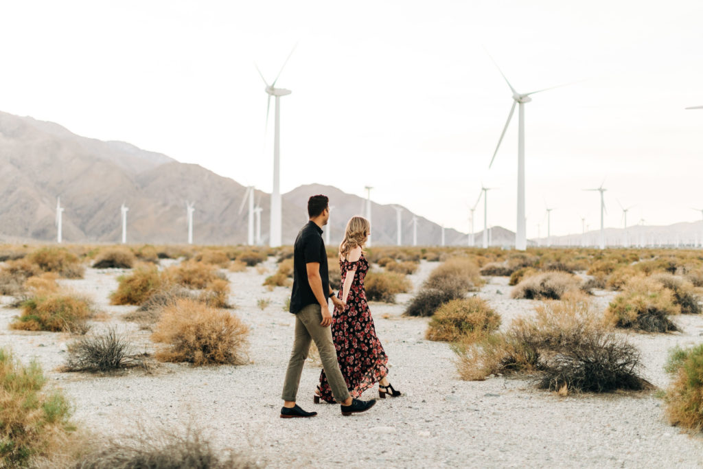 southern california engagement photos; couple walking with windmills in the background in palm springs