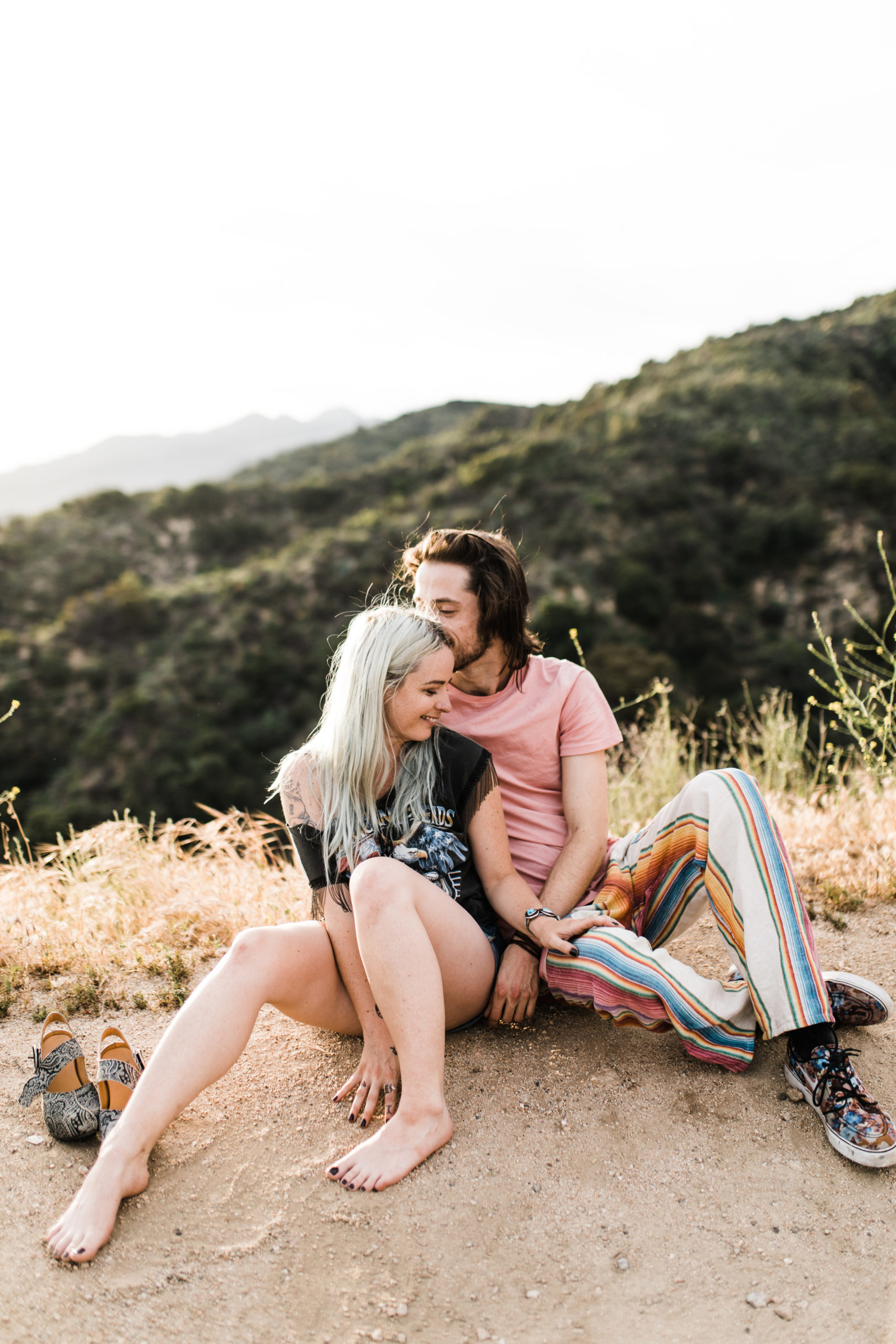 southern california engagement photos locations; couple sitting on the ground laughing while the man kisses the woman on the temple
