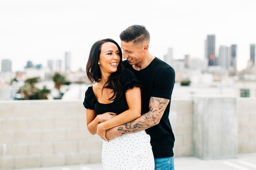 rooftop engagement photos in los angeles; a couple smiling and embracing on a rooftop with buildings in the background in los angeles, ca