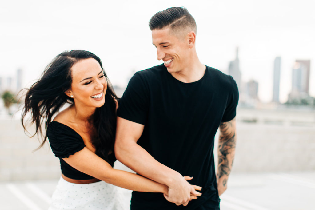 rooftop engagement photos in los angeles; a couple smiling and embracing on a rooftop with buildings in the background in los angeles, ca