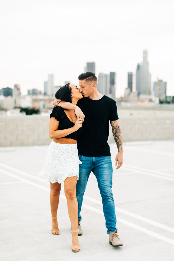 rooftop engagement photos in los angeles; a couple kissing and walking on a rooftop with buildings in the background in los angeles, ca
