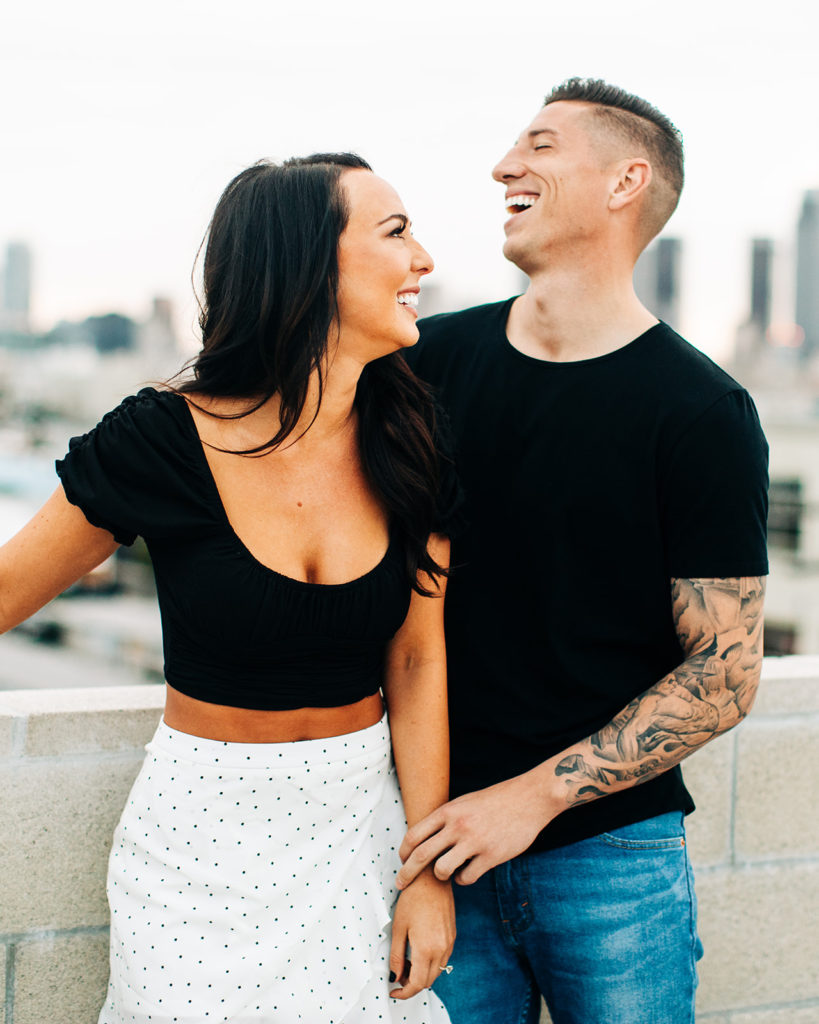 rooftop engagement photos in los angeles; a couple smiling and laughing on a rooftop with buildings in the background in los angeles, ca