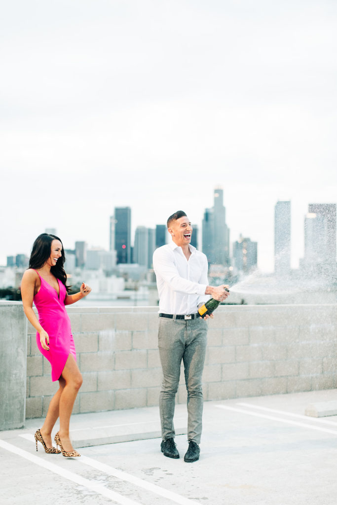 rooftop engagement photos in los angeles; a couple in formal outfits popping champagne on a rooftop with buildings in the background in los angeles, ca