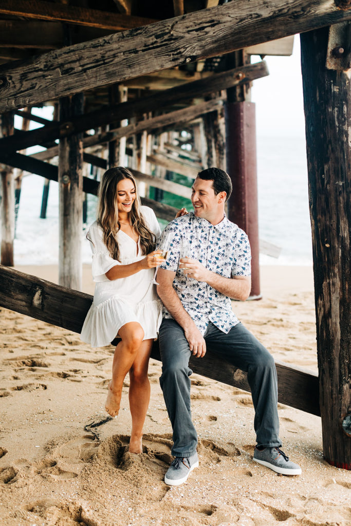 southern california engagement photos; man and woman cheers their champagne glasses while sitting at the beach