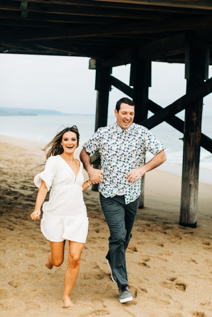 southern california engagement photos; man and woman running while holding hands at the beach