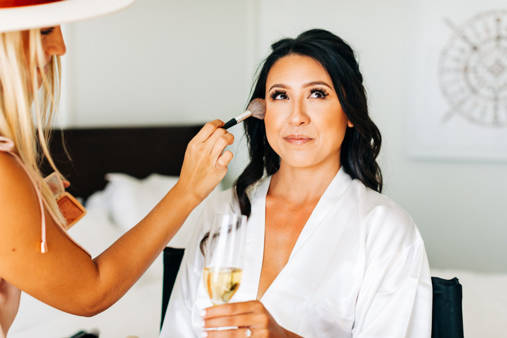 Crystal Cove Micro Wedding in Orange County; bride getting her makeup done on her wedding day while holding champagne