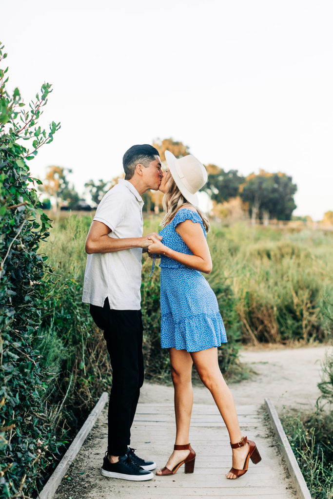 Newport Back Bay Engagement Photos; man and woman couple kissing while surrounded by greenery. the woman has on a blue dress with white dots and the man has on a gray collared shirt with black pants.