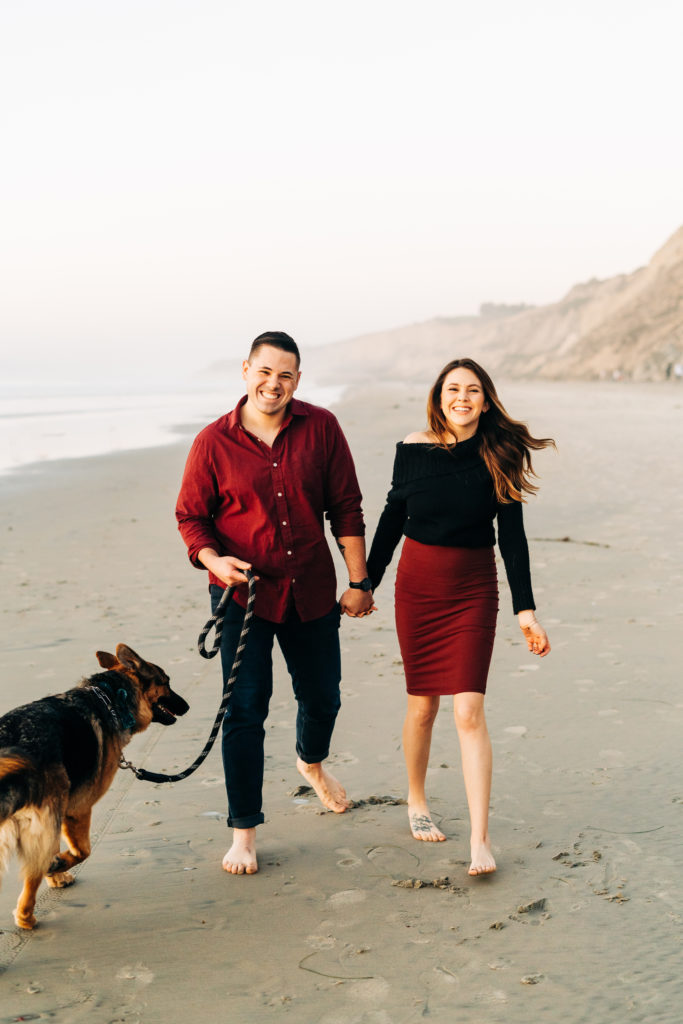 black's beach engagement photos; san diego wedding photographer; a couple holding hands and walking on the beach with their dog, they are smiling at the camera