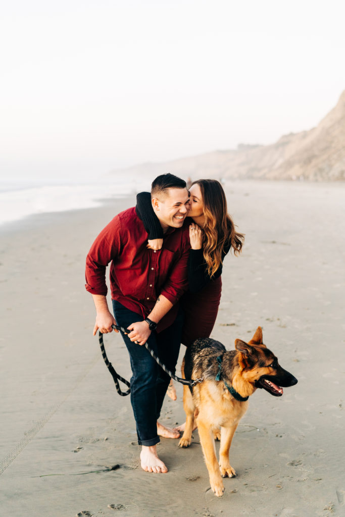 black's beach engagement photos; san diego wedding photographer; a woman jumps on the back of her husband, they are at the beach with their dog