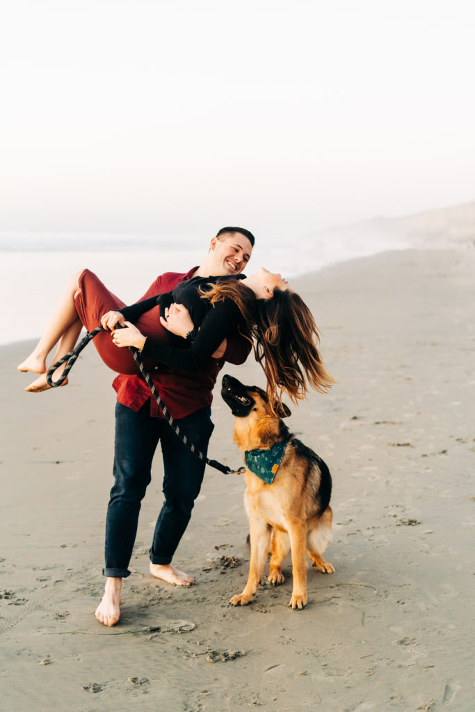 black's beach engagement photos; san diego wedding photographer; husband picks up his wife and spins her around, they are at the beach with their dog