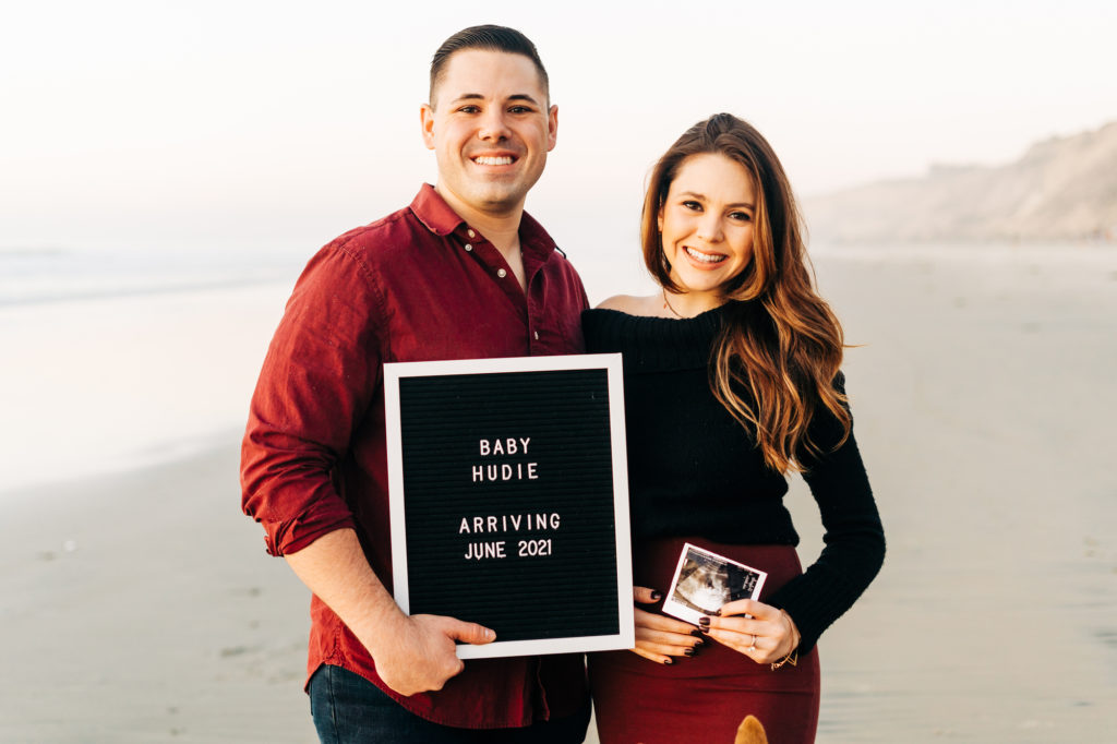 black's beach engagement photos; san diego wedding photographer; a couple holding a sign that says "baby hudie arriving june 2021", the woman is holding a sonogram