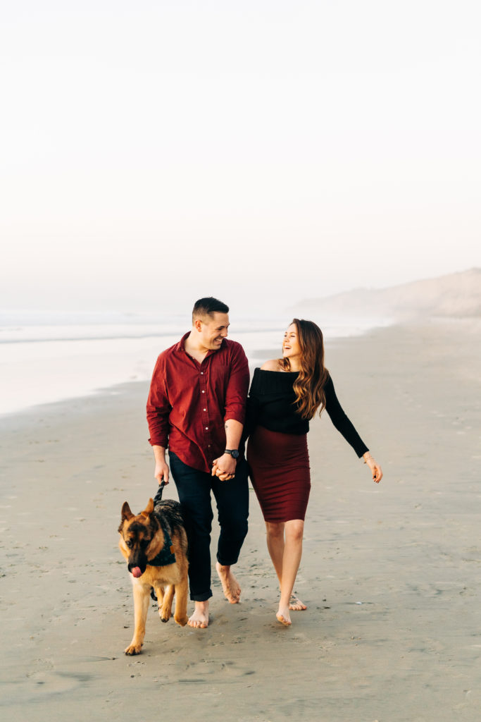 black's beach engagement photos; san diego wedding photographer; a couple walking with their dog on the beach, they are smiling at each other