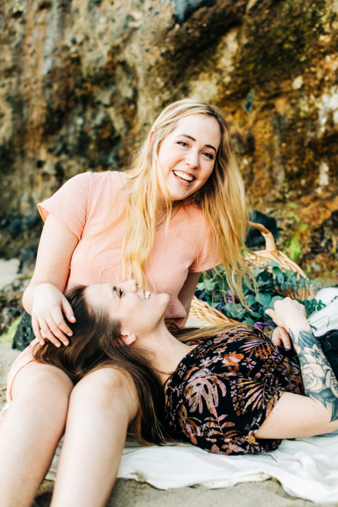 LGBTQ+ wedding photographer in san diego; a lesbian couple having a picnic at the beach, one is laying down in the other's lap, one is smiling at the camera