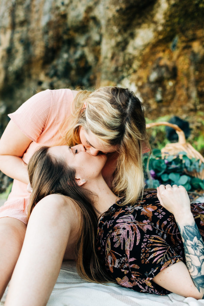 LGBTQ+ wedding photographer in san diego; a lesbian couple kissing each other, one is lying in the lap of the other, the are sitting on the sand at the beach