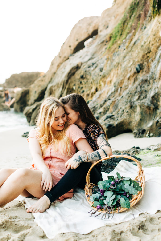 LGBTQ+ wedding photographer in san diego; a lesbian couple having a picnic at the beach, they are both smiling and one is about to kiss the other on the cheek