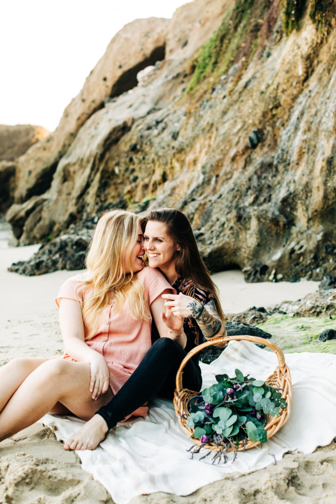 LGBTQ+ wedding photographer in san diego; a lesbian couple sitting on the beach having a picnic, they are sitting close together and laughing