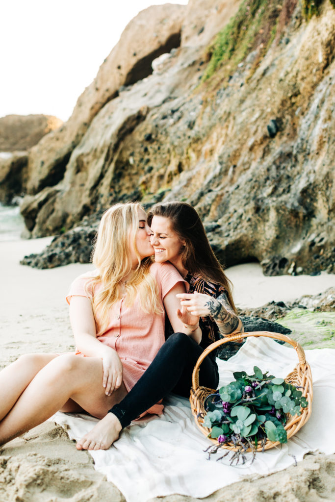 LGBTQ+ wedding photographer in san diego; a lesbian couple sitting on the sand at the beach having a picnic, one is kissing the other on the cheek