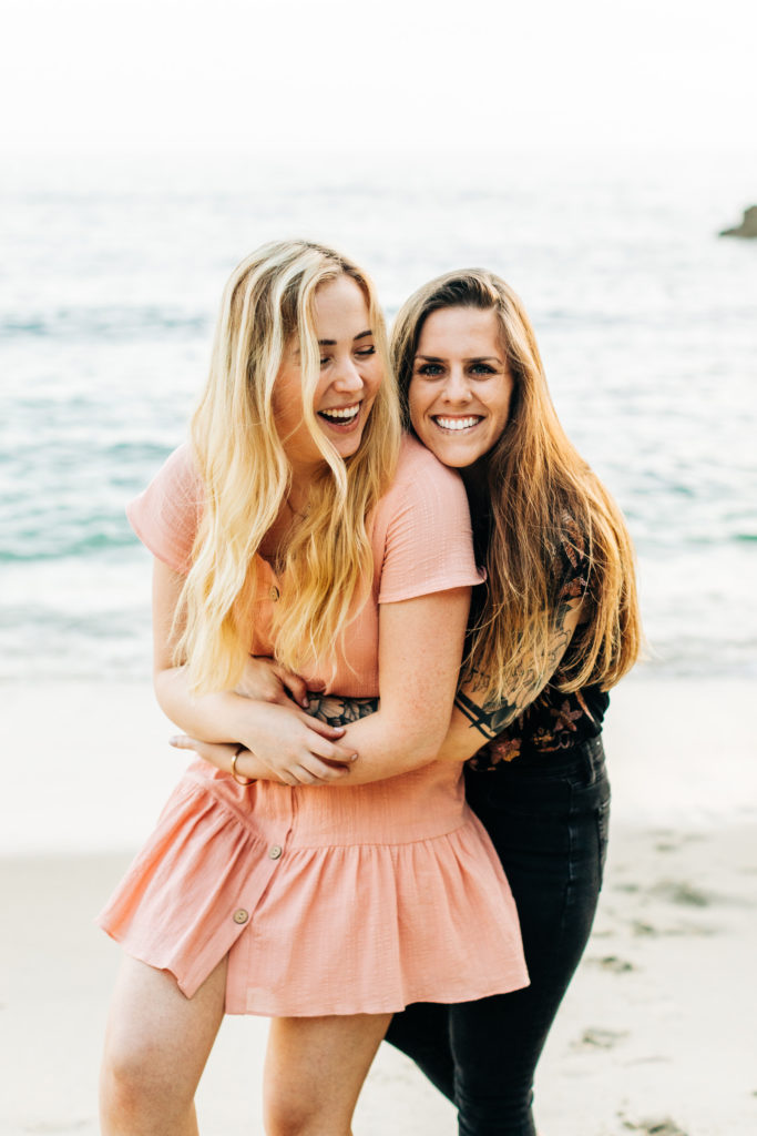 LGBTQ+ wedding photographer in san diego; a woman hugs her girlfriend from behind, they are smiling, they are at the beach