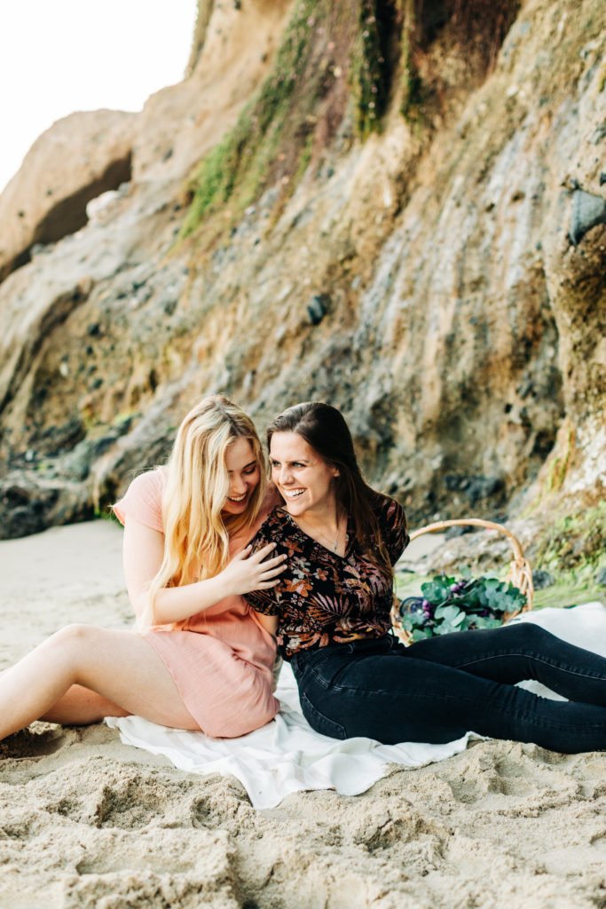 LGBTQ+ wedding photographer in san diego; a lesbian couple sitting next to each other on the beach, they are laughing