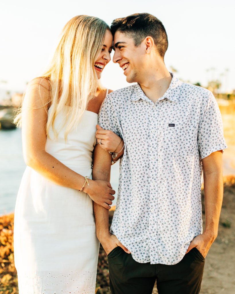 san diego engagement photos at sunset cliffs; san diego wedding photographer; a woman and man holding each other, they are smiling, the beach and sunset cliffs in san diego is in the background