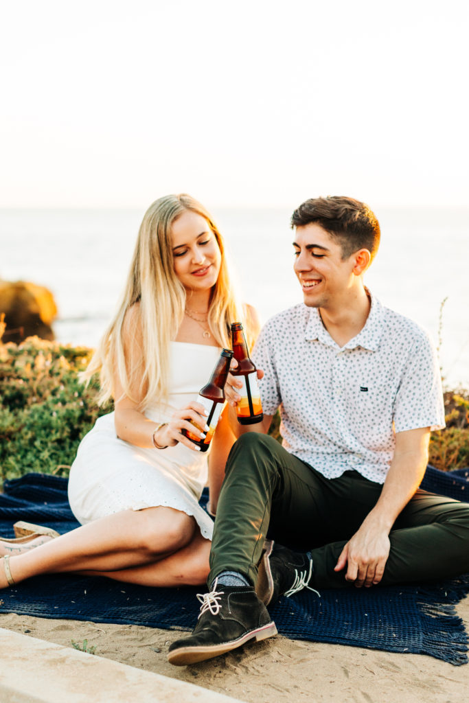 san diego engagement photos at sunset cliffs; san diego wedding photographer; a man and woman cheers their beer bottles together