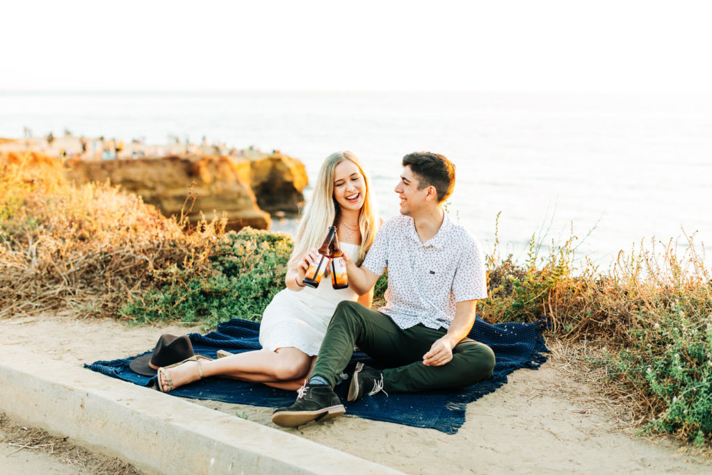 san diego engagement photos at sunset cliffs; san diego wedding photographer; a man and woman cheers their beer bottles together, they are sitting on a blanket overlooking the ocean at sunset cliffs in san diego