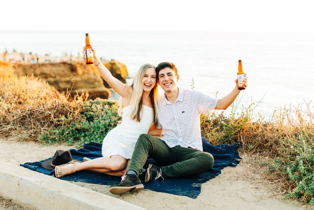 san diego engagement photos at sunset cliffs; san diego wedding photographer; a man and woman couple sitting on a blanket at sunset cliffs in san diego, they are holding beer bottles up and smiling