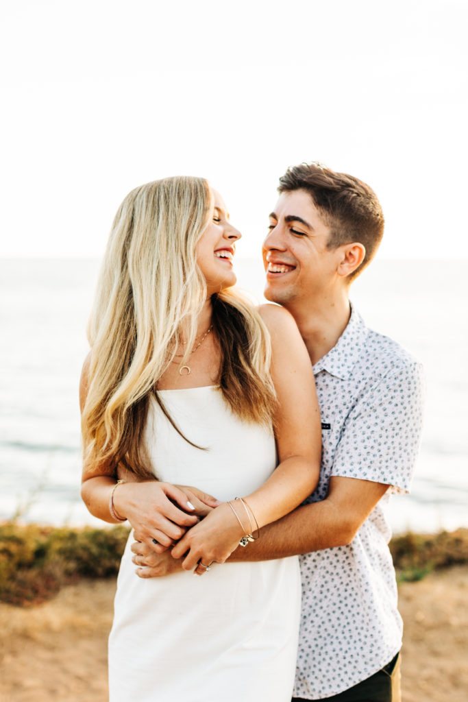 san diego engagement photos at sunset cliffs; san diego wedding photographer; a man hugs his girlfriend from behind, they are smiling