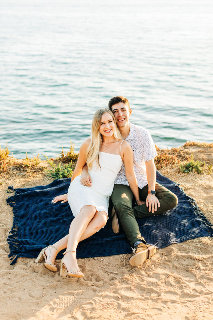 san diego engagement photos at sunset cliffs; san diego wedding photographer; a man and woman sitting on a blanket on a cliff overlooking the ocean, they are smiling at the camera