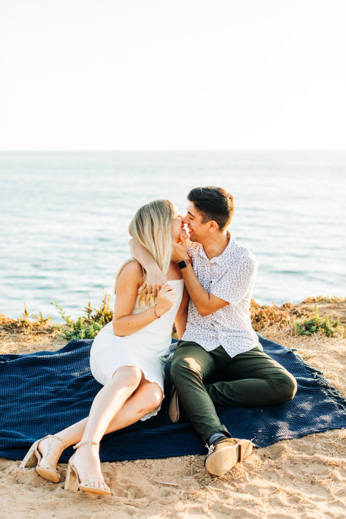 san diego engagement photos at sunset cliffs; san diego wedding photographer; a man and woman sitting on a blanket on a cliff overlooking the ocean, they are about to kiss