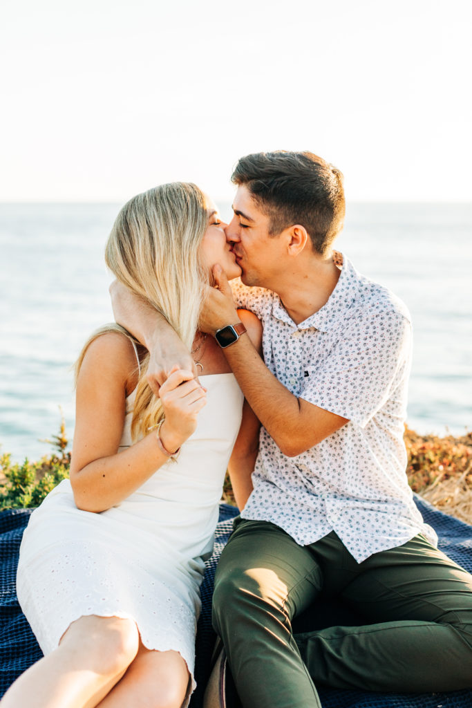 san diego engagement photos at sunset cliffs; san diego wedding photographer; a man and woman kissing, they are sitting on a blanket on a cliff overlooking the ocean