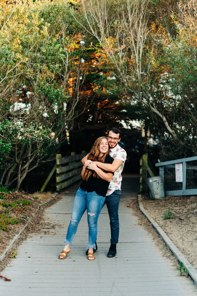 Sunset Engagement Photos at Morro Bay, morro bay wedding photographer; a man hugs his girlfriend from behind, they are in front of some trees on a cement pathway