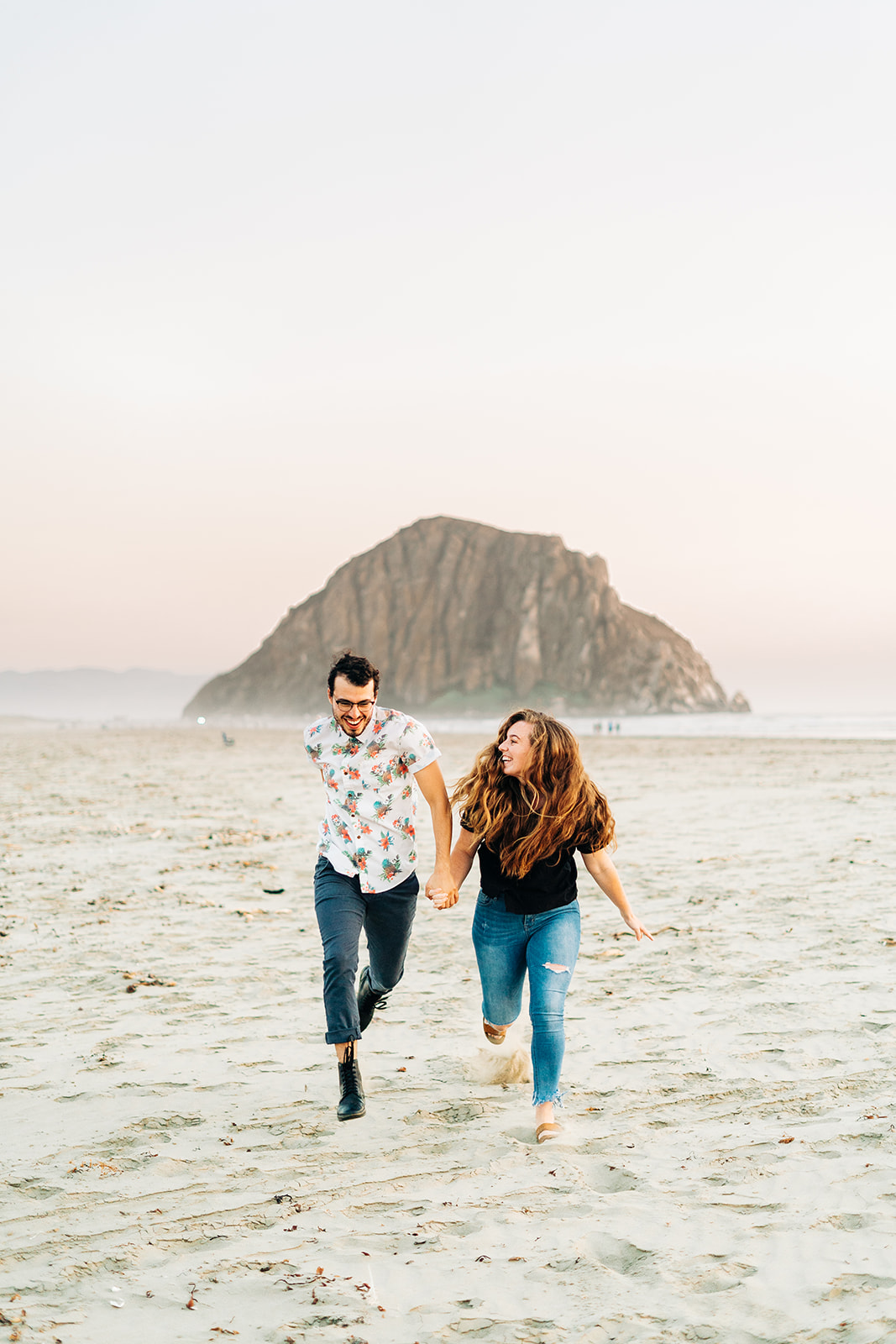 Sunset Engagement Photos at Morro Bay, morro bay wedding photographer; a man and woman holding hands running towards the camera, they are running on the sand and the Morro Bay rock formation is behind them.