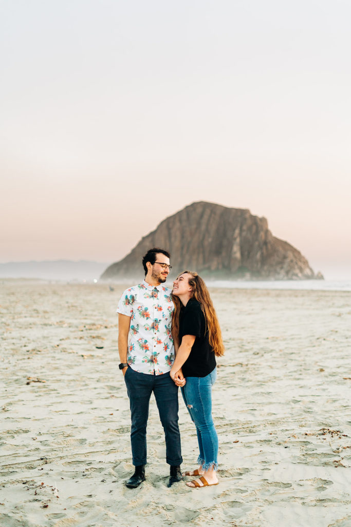 Sunset Engagement Photos at Morro Bay, morro bay wedding photographer; a man and woman couple looking at each other and smiling at the beach in Morro Bay, California