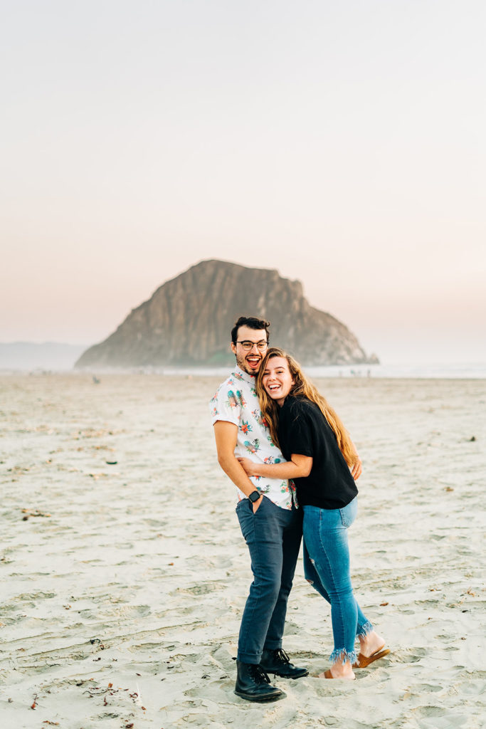 Sunset Engagement Photos at Morro Bay, morro bay wedding photographer; a man and woman laughing at the camera while they hug each other in morro bay, california
